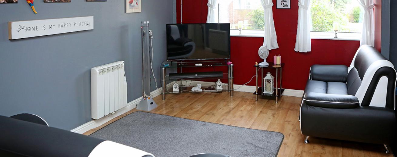 Grey and deep red seating area with wooden flooring and two blank and white leather sofas, a glass television unit with matching stand hosting homely candles and soft lighting for the room.