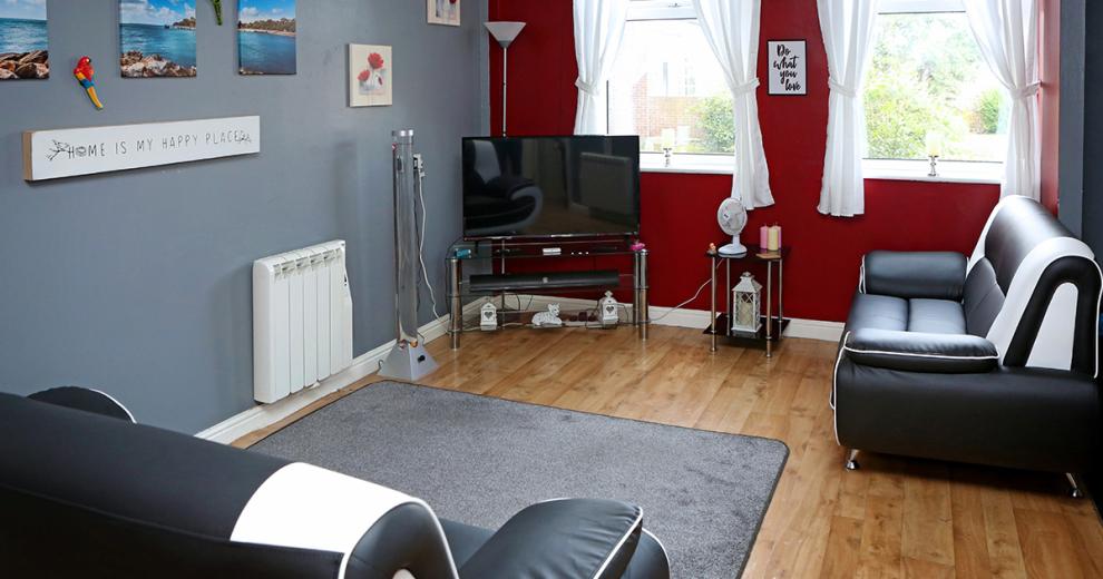 Grey and deep red seating area with wooden flooring and two blank and white leather sofas, a glass television unit with matching stand hosting homely candles and soft lighting for the room.