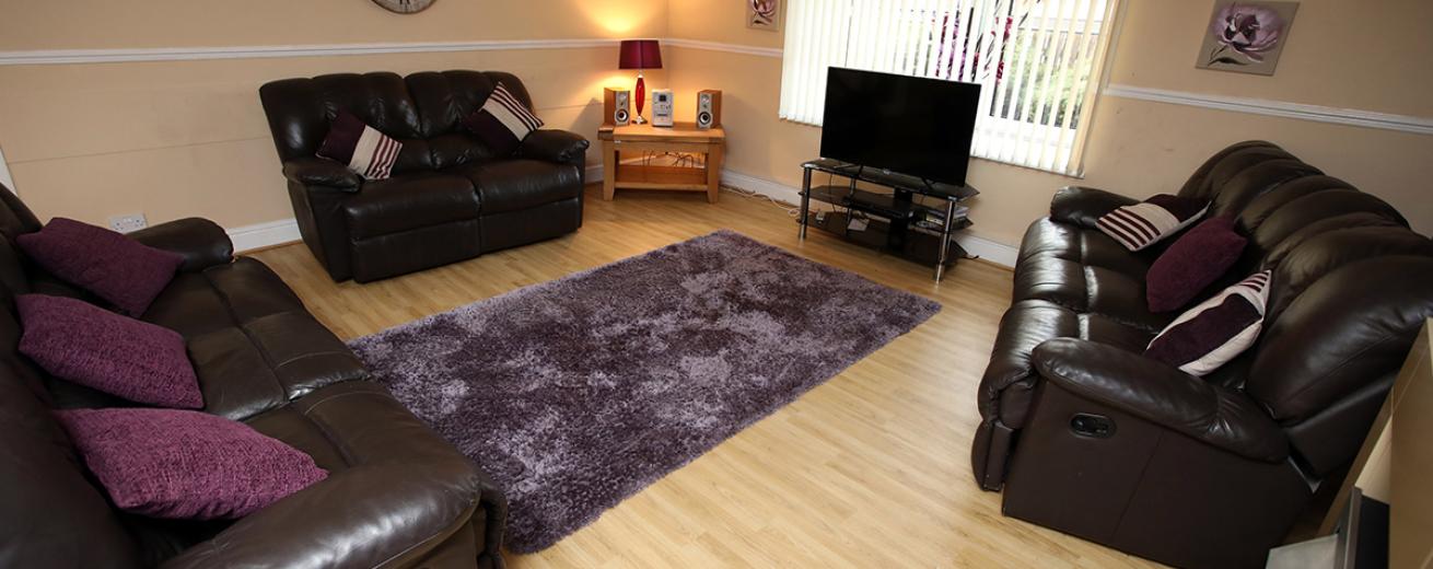 Comfortable, softly lit living area with a mauve rug central to the room which matches the wall art and lamp decorations, surrounded by two three-seater leather sofas and one two seater leather sofa all facing towards a television resting on a black glass unit.