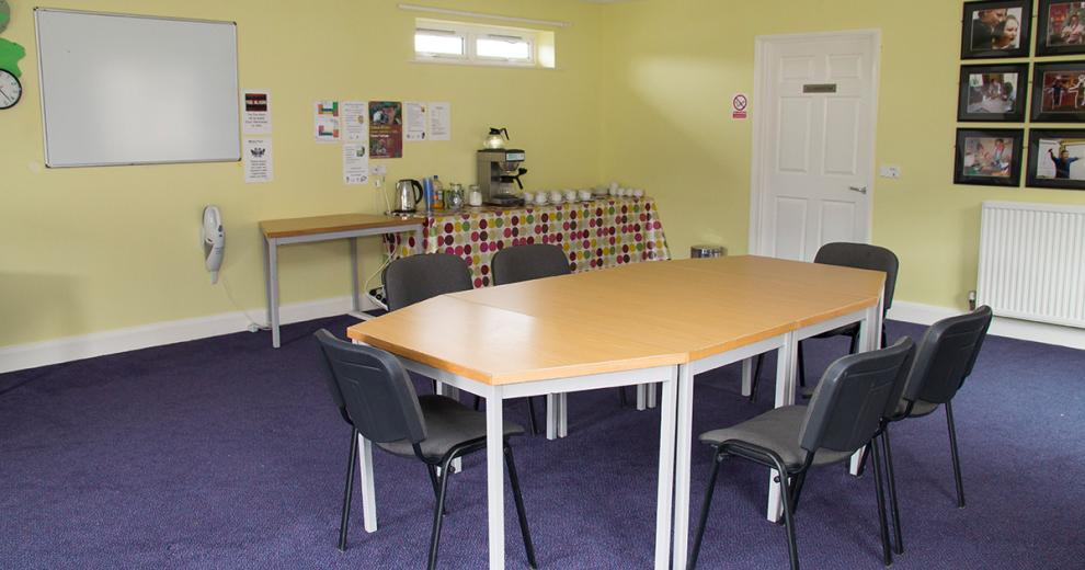 Bold yellow painted room with purple carpet to give personality to a simplistic room which has a wall mounted white board and large octagonal table central to the room. The tea making facilities offer a kettle and coffee maker an array of accompaniments on a spotty table covering.