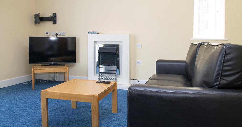 A minimal relaxing cream and blue seating room with wall mounted silver electric fire, three seater leather sofa facing a television placed on one of two wooden coffee tables.