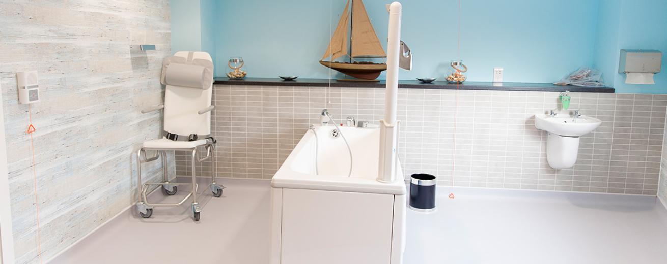 Nautical themed wetroom, consisting of blue painted feature wall, mottled wall tiles, model ship and decorative seashells on the shelf to create a tranquil space to relax in the bath or shower whilst having assistance cords around the room and shower chair available.