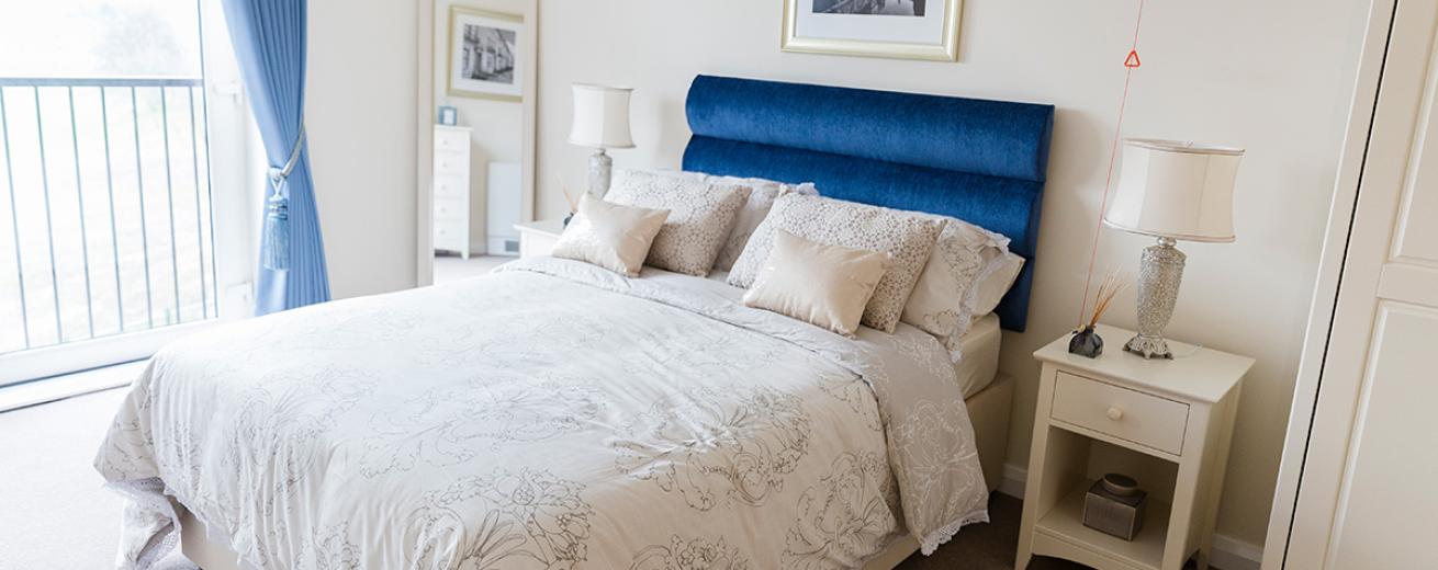 Luxurious double bedroom using pops of blue colouring in the velvet headboard and curtains whilst keeping the wardrobe, bedside table and floor length mirror all a white wood. The airiness to the room comes from the sliding window leading onto the balcony.