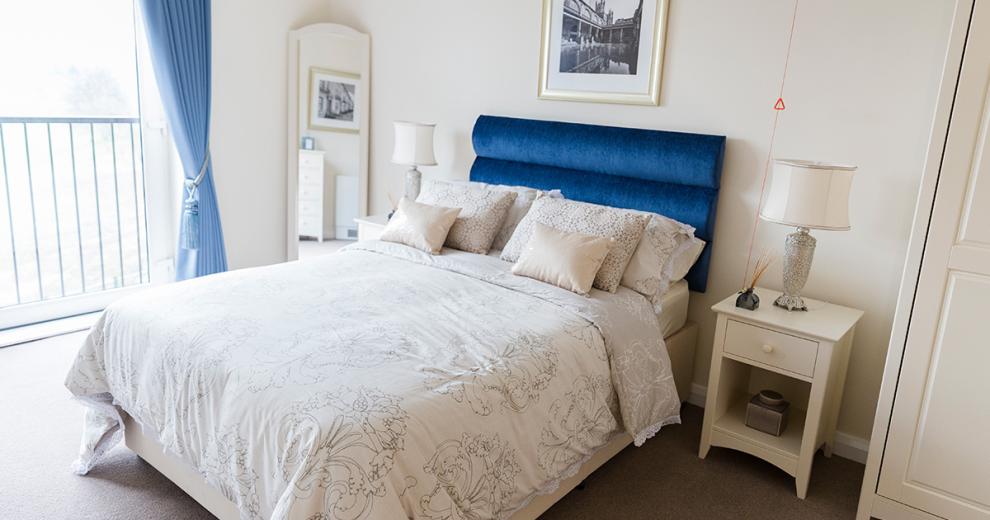 Luxurious double bedroom using pops of blue colouring in the velvet headboard and curtains whilst keeping the wardrobe, bedside table and floor length mirror all a white wood. The airiness to the room comes from the sliding window leading onto the balcony.