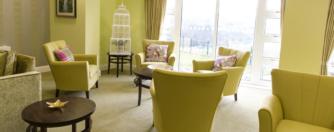 Tasteful lime green armchairs with purple, green and silver bird cushions surround black circular coffee tables, with a cream carpet to soften the colourful furniture the room is flooded with natural light from the floor to ceiling windows and doors leading out into the garden.