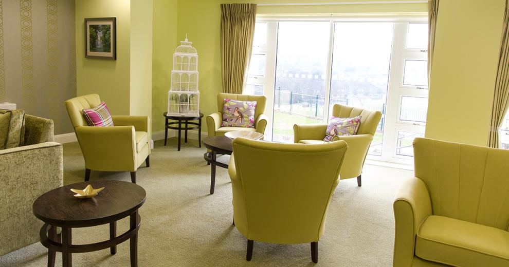 Tasteful lime green armchairs with purple, green and silver bird cushions surround black circular coffee tables, with a cream carpet to soften the colourful furniture the room is flooded with natural light from the floor to ceiling windows and doors leading out into the garden.