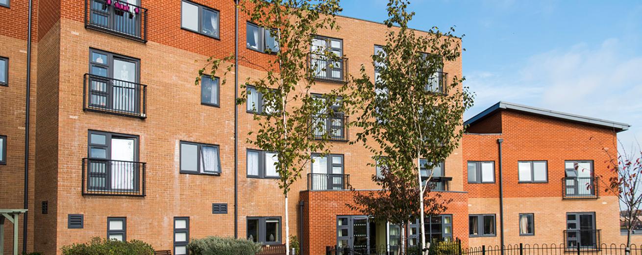 Beautiful contemporary apartment building contrasting dark grey window frames with the multitoned red bricked exterior. The outdoor area offers metal seating as well as numerous wooden benches all looking out to the newly planted trees and garden area.