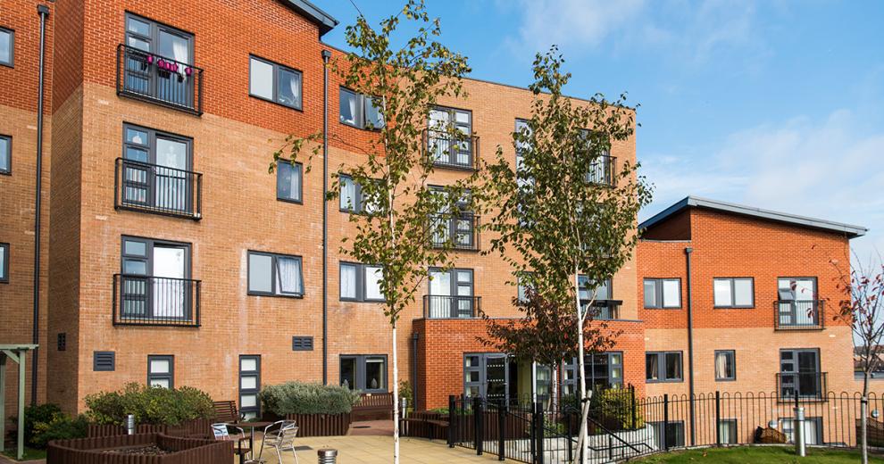 Beautiful contemporary apartment building contrasting dark grey window frames with the multitoned red bricked exterior. The outdoor area offers metal seating as well as numerous wooden benches all looking out to the newly planted trees and garden area.