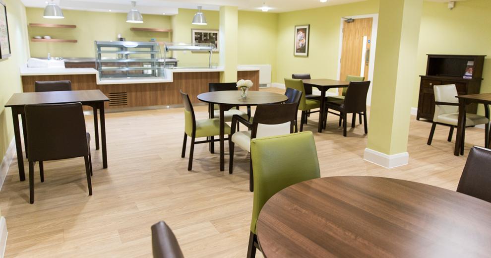 Soft green restaurant area with orderly hot counter and an array of mixed sized dining tables offering between two- and four-seater options. A mixture of square and circular dark wooden tables matched with green, cream, and brown seating surrounding them.