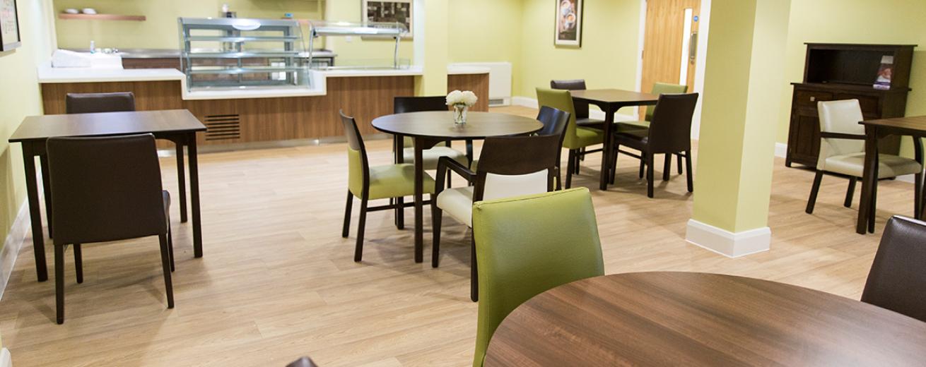 Soft green restaurant area with orderly hot counter and an array of mixed sized dining tables offering between two- and four-seater options. A mixture of square and circular dark wooden tables matched with green, cream, and brown seating surrounding them.