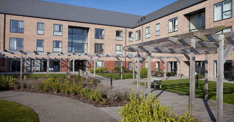 Stylized retirement living community with an array of large windows over looking a sectioned off grassed areas, flower beds and walkways, located in Stoke-on-Trent.