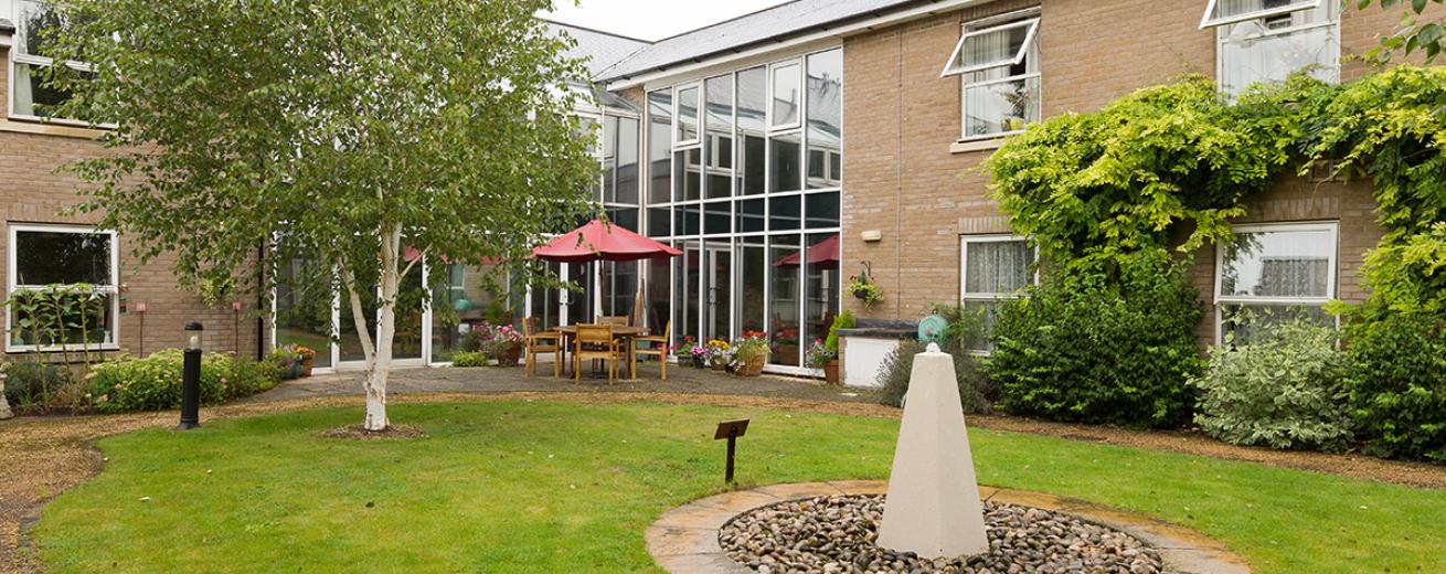 Tranquil outdoor area circulating around a simplistic water feature in the centre of the grass. A four-person wooden dining table and chairs is topped by a red parasol, located just in front of the glass window panels spanning the corner of the building. A healthy amount of low maintenance vegetation is flourishing around the boarder of the garden.