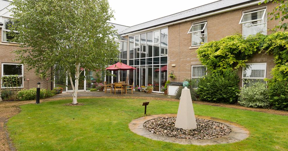 Tranquil outdoor area circulating around a simplistic water feature in the centre of the grass. A four-person wooden dining table and chairs is topped by a red parasol, located just in front of the glass window panels spanning the corner of the building. A healthy amount of low maintenance vegetation is flourishing around the boarder of the garden.