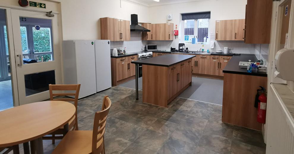A generious sized kitchen and dining area, with wooden cabinetry, black worktops a static floating island in the centre. Two large fridge freezers sit adjacent to the doorway leading outside. Two ovens and hobs sit next to each other and multiple tea making facilities are dotted around the work surface.