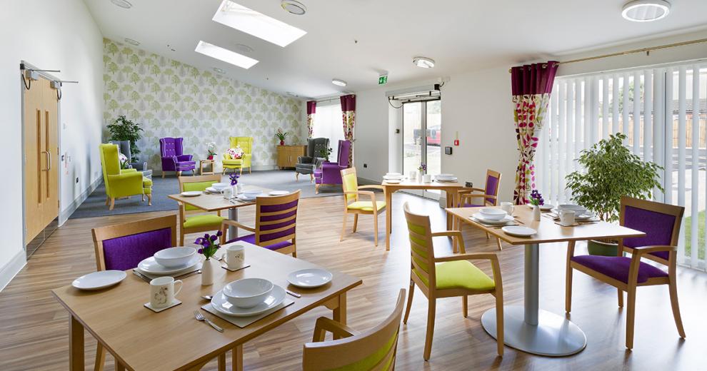 Sun lit restaurant area filled with four two seated wooden squared tables and contrasting lime green and deep purple chairs.