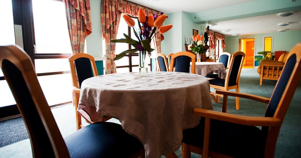 A bright inviting dining area decorated in pastel green walls and matching carpets with multiple circular tables draped in cream patterned tablecloths. The navy upholstery on the chairs compliments the orange floral decorations on the tables and curtains.