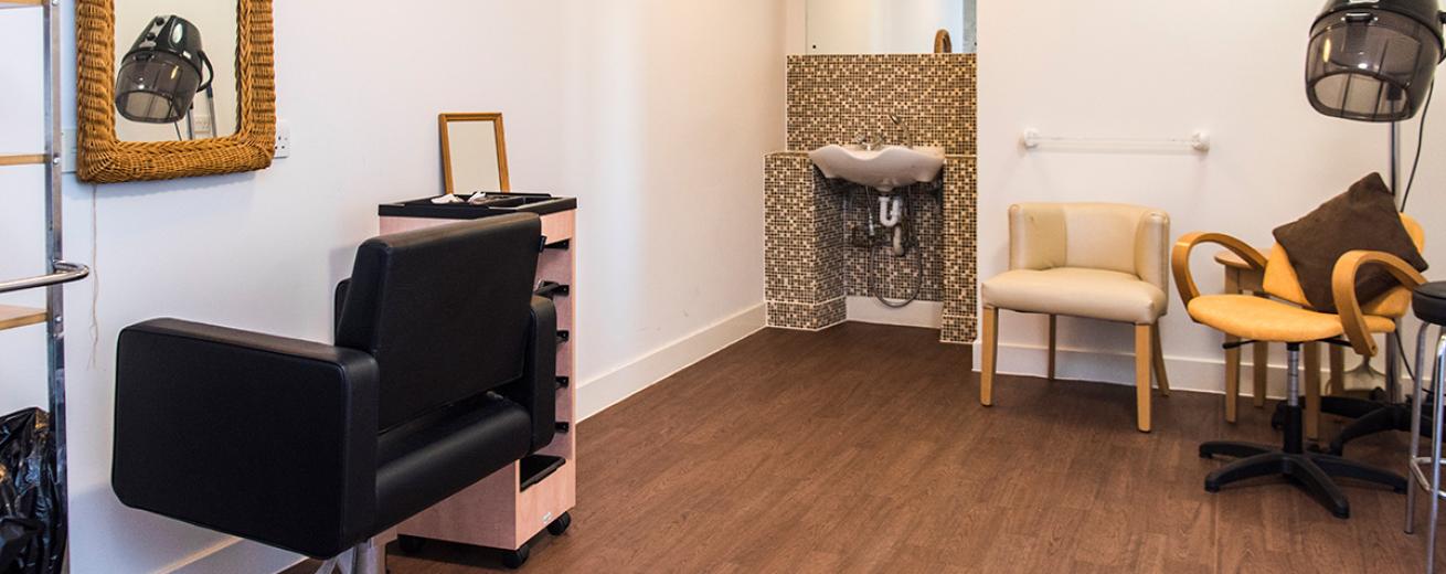 Simplistic and clean hair salon area. With a bold mosaic decorated alcove with hair washing basin tucked away from the styling area. A plush black styling chair is located in front of a wicker framed mirror. A yellow styling chair sits underneath an industrial hairdryer.