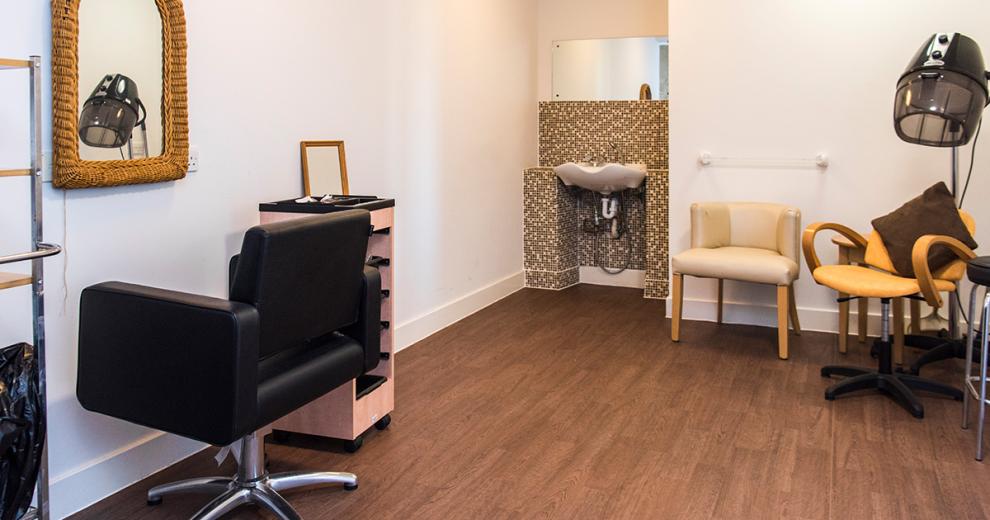 Simplistic and clean hair salon area. With a bold mosaic decorated alcove with hair washing basin tucked away from the styling area. A plush black styling chair is located in front of a wicker framed mirror. A yellow styling chair sits underneath an industrial hairdryer.