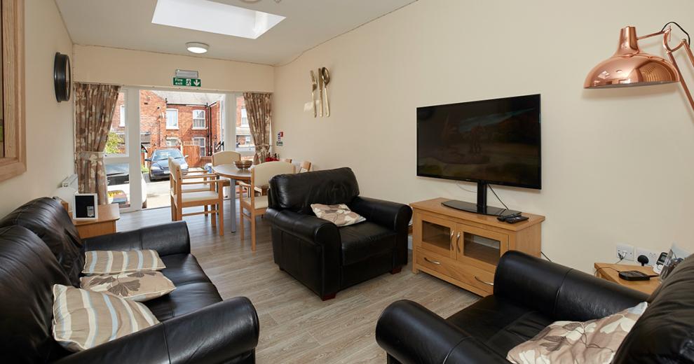 Snug seating area consisting of two-seater, black leather sofa, and two matching armchairs surrounding a television on a small television unit and rose gold floor lamp.