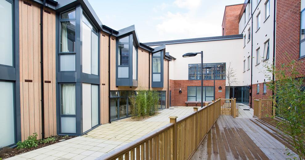 Contemporary wood clad building with large glass window panels, joining on to a brick and rendered four storey building, with central patio and decking at Defoe Court.