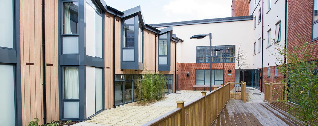 Contemporary wood clad building with large glass window panels, joining on to a brick and rendered four storey building, with central patio and decking at Defoe Court.