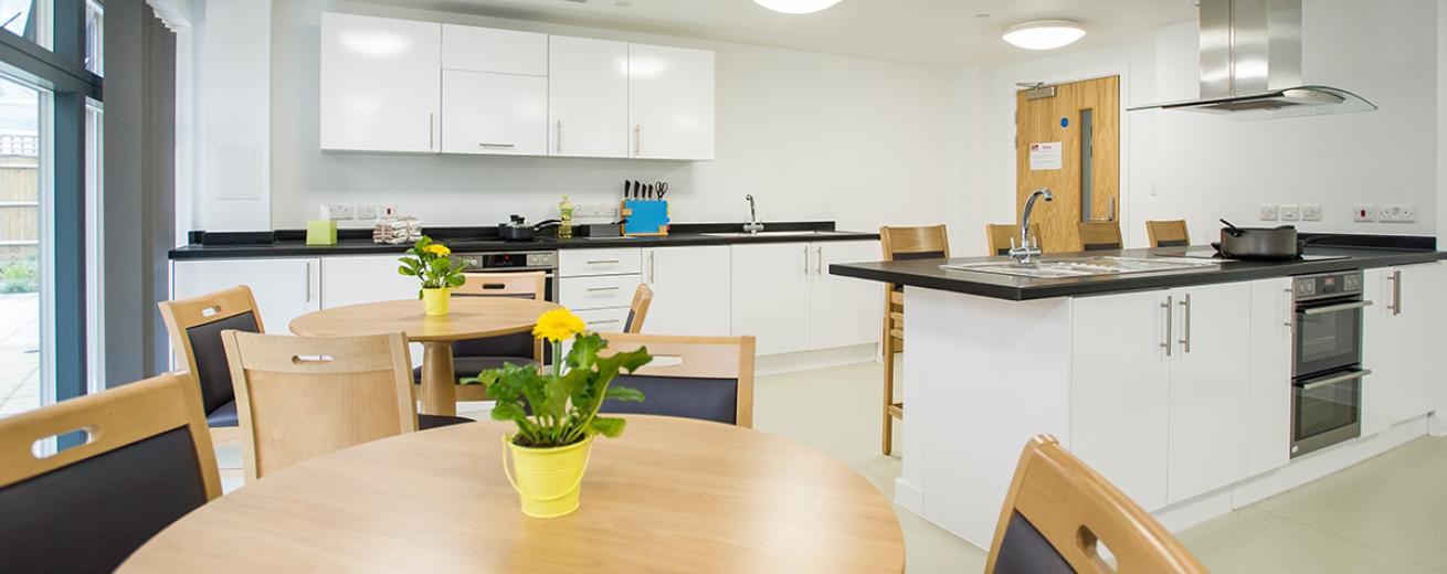 Two generous sized kitchen units both consisting of multiple storage cupboards, oven, hob and sinks. One unit has four breakfast bar seating, as well as two circular dining table with potted plants next to floor to ceiling windows onto a patio area.