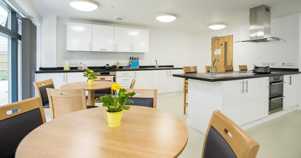 Two generous sized kitchen units both consisting of multiple storage cupboards, oven, hob and sinks. One unit has four breakfast bar seating, as well as two circular dining table with potted plants next to floor to ceiling windows onto a patio area.