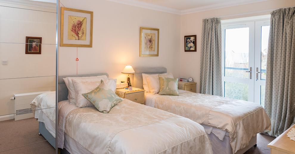 A beautifully airy twin bedroom, featuring two light grey divan beds decorated in satin bedding. A pair of bedside tables nestle between the beds, a large pair of french doors allow a mass amount of natural light.
