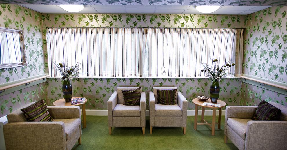 A bold green and white floral-patterned wallpaper creates a cosier feel to the small seating area. The green carpet compliments the decoration and simple neutral armchairs, and small circular coffee tables soften the look of the room.