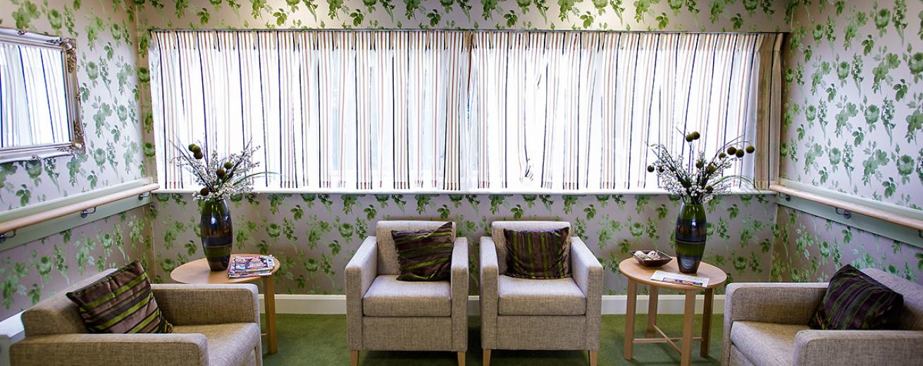 A bold green and white floral-patterned wallpaper creates a cosier feel to the small seating area. The green carpet compliments the decoration and simple neutral armchairs, and small circular coffee tables soften the look of the room.