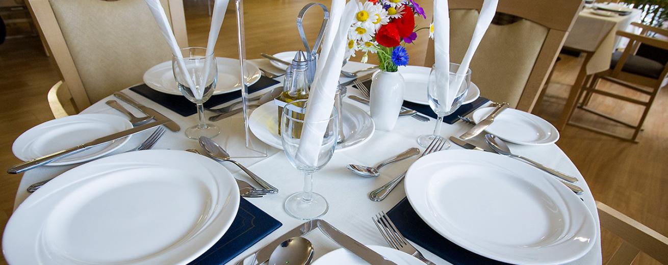 A beautifully presented dining table. Covered in a white pressed linen cloth, black mats, dining plates and side plates. Cutlery set for a three course dining experience and wine glasses with napkins.