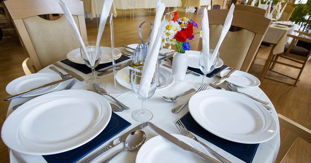 A beautifully presented dining table. Covered in a white pressed linen cloth, black mats, dining plates and side plates. Cutlery set for a three course dining experience and wine glasses with napkins.