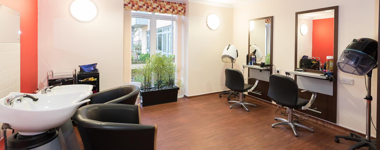 A large hairdressing salon with a vivid red feature wall, behind the two white hair washing basins. A professional set up salon with wall mounted industry style mirrors, chairs and two portable hair dryers.