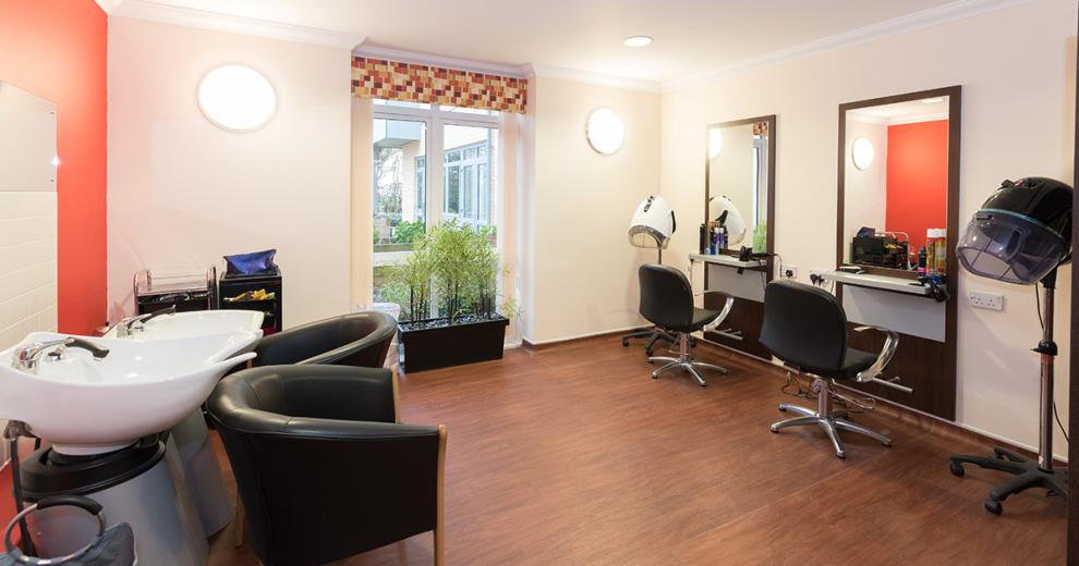 A large hairdressing salon with a vivid red feature wall, behind the two white hair washing basins. A professional set up salon with wall mounted industry style mirrors, chairs and two portable hair dryers.