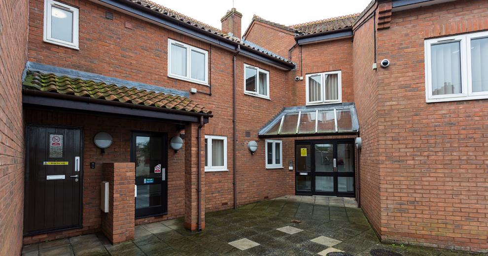 Multi-entranced brick exterior with terracotta clay roof tiles. Multiple security cameras and lighting. Accessed by a clean concrete tiled walkway to Drawbridge House.