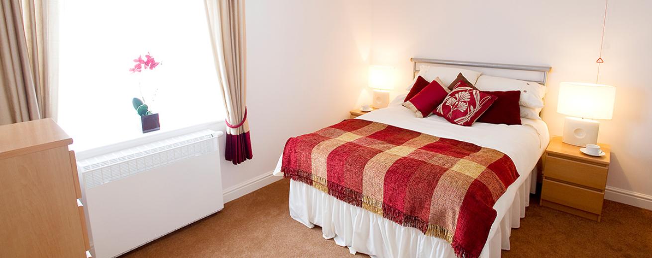 A cosy double bedroom neutrally decorated with homely red finishing touches. The room has matching light wooden furniture set consisting of a large chest of draws and two bedside tables with matching white lamps.