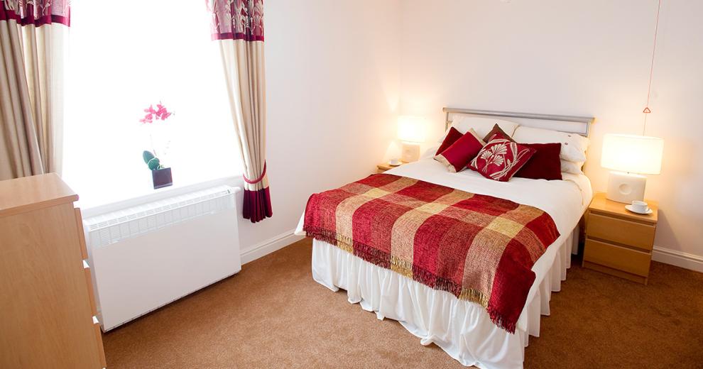 A cosy double bedroom neutrally decorated with homely red finishing touches. The room has matching light wooden furniture set consisting of a large chest of draws and two bedside tables with matching white lamps.