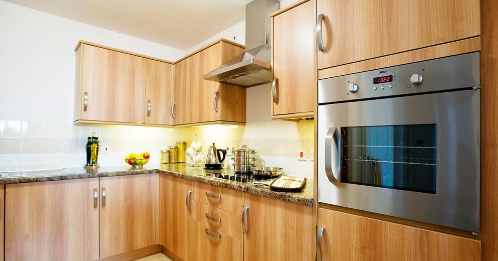 A large domestic kitchen featuring wall mounted cabinetry and draws. With a fitted ceramic hob centred on the worktop next to a wall mounted oven. The marble effect counter pulls the colouring from the hob, tiles and cabinets.