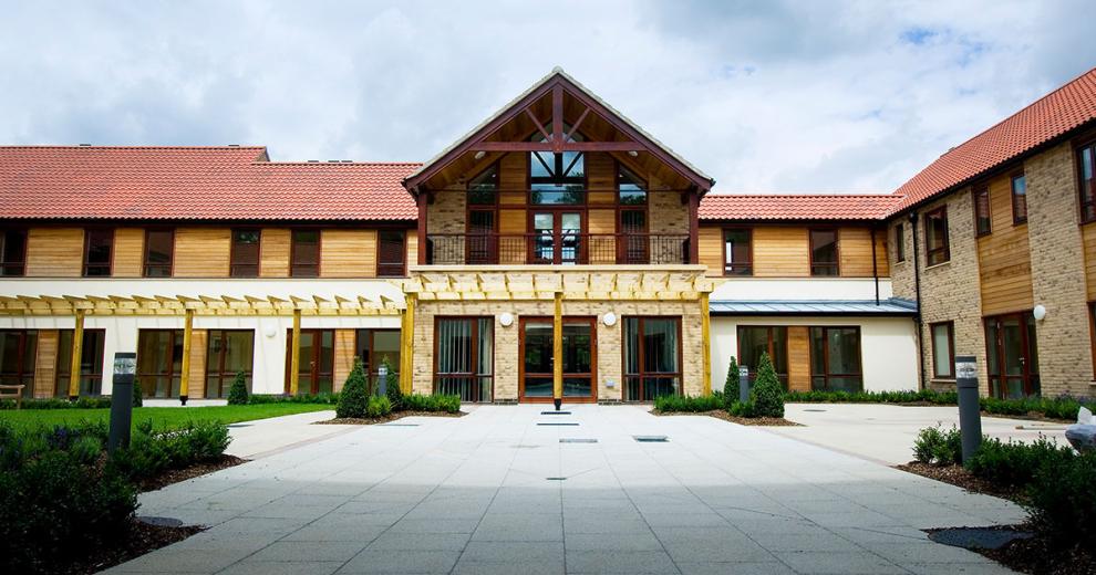 A luxurious property with a grand entrance and first storey balcony overlooking the white slabbed walkway. The property has been built using a mixture of natural materials such as brick, wooden panelling, and wooden beams. Small shrubbery plants line the perimeter.
