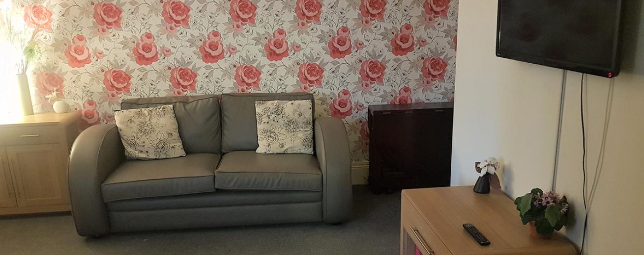 Bold red floral feature wall used in the communal seating area, in front sits a grey curved armed leather sofa and wooden side units. A wall mounted television is mounted on the white painted wall withy a small television unit underneath.