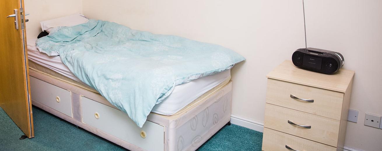 A minimalistic single bed room. The divan bed tucks neatly behind the door and into the corner of the room. A small three draw wooden chest of draws with a radio sat ontop. The teal carpets add a pop of colour to the space.