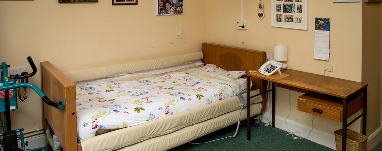 An assisted living bed, tucked neatly into the corner of the room with plenty of room to manoeuvre in and out safely, next to the bed is a wooden desk and the walls are adorned with personal photographs.