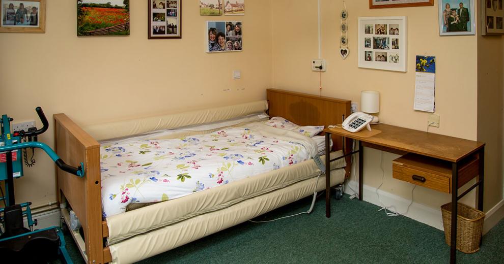 An assisted living bed, tucked neatly into the corner of the room with plenty of room to manoeuvre in and out safely, next to the bed is a wooden desk and the walls are adorned with personal photographs.