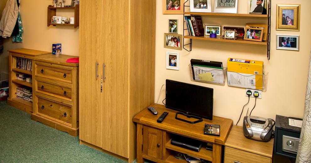 Mass amounts of wooden furniture line the largest wall in the room nestled next to each other are a small oak bookshelf, three draw large chest of draws, a single wardrobe, a television unit and small television another three draw unit and mass amounts of personal photographs and documentation safely tucked away but easily accessible on the walls.