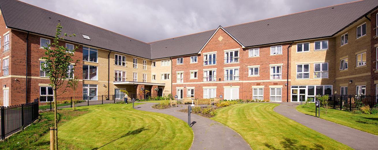 The newly built, yellow, and red bricked, three storey retirement living property set behind winding pathways which lead to a circular outdoor seating area as well the property and lovely grassed areas.