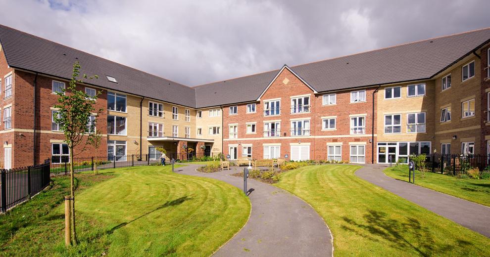 The newly built, yellow, and red bricked, three storey retirement living property set behind winding pathways which lead to a circular outdoor seating area as well the property and lovely grassed areas.