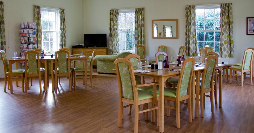 A spacious wooden laminate floored dining area, the room has an array of light wooden tables, matching chairs with a green and white floral upholstered coverings to add some colour and brightness to the room. The multi green and white vector leaf patterned curtains tie the colour scheme in.