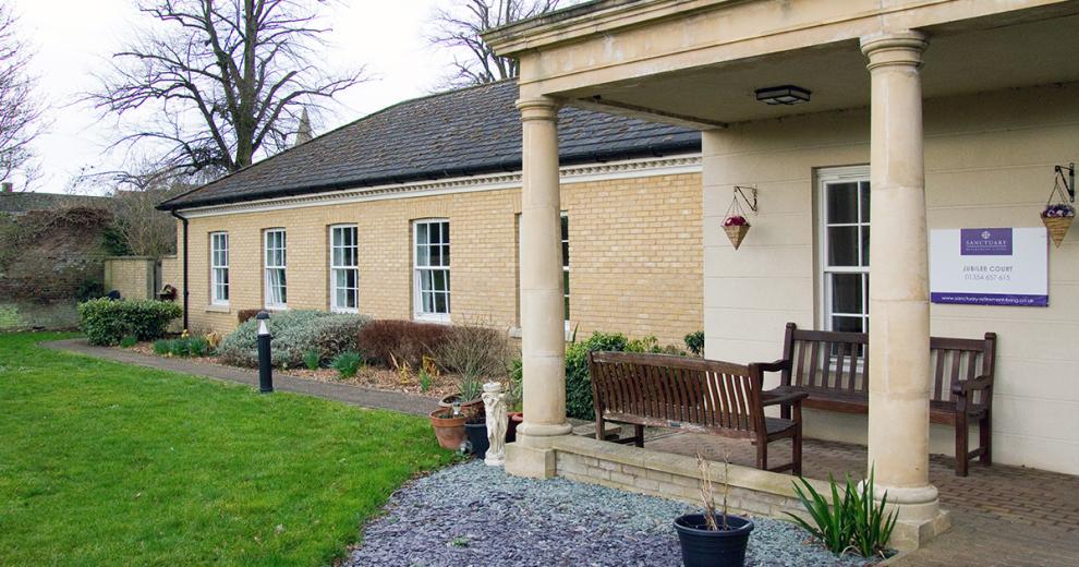 A period designed stone built building is home to the entrance of the property, the entrance is covered by a stone built porch with chunky columns supporting the roof. Two wooden benches sit underneath looking out onto the gardens. The rest of the building is built with soft coloured yellow bricks and long rectangular windows.
