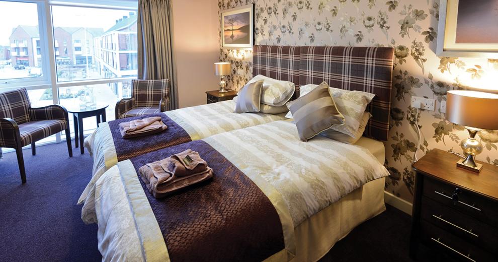 A cosy atmospheric bedroom area using dark colour tones with a deep purple carpet and a mixtures of purples for a checked headboard. The two fabric chairs also match the checked headboard. The room is decorated with multitoned  green peonies repeated all over the back feature wall. Beside the two single beds pushed together are two three drawer bedside tables each with a small lamp. A massive window fills the wall space overlooking the courtyard.