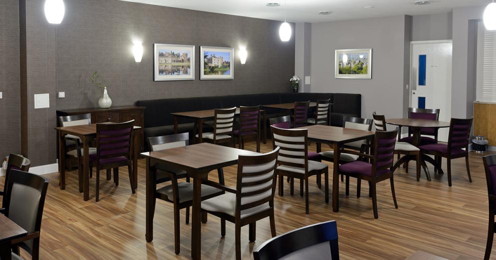 The dining room has a monochrome palette with the walls being painted in different grey tones with silver picture frames hanging on them. The multiple wooden dining tables are stained a dark mahogany with light grey and deep aubergine coloured cushioning to them.